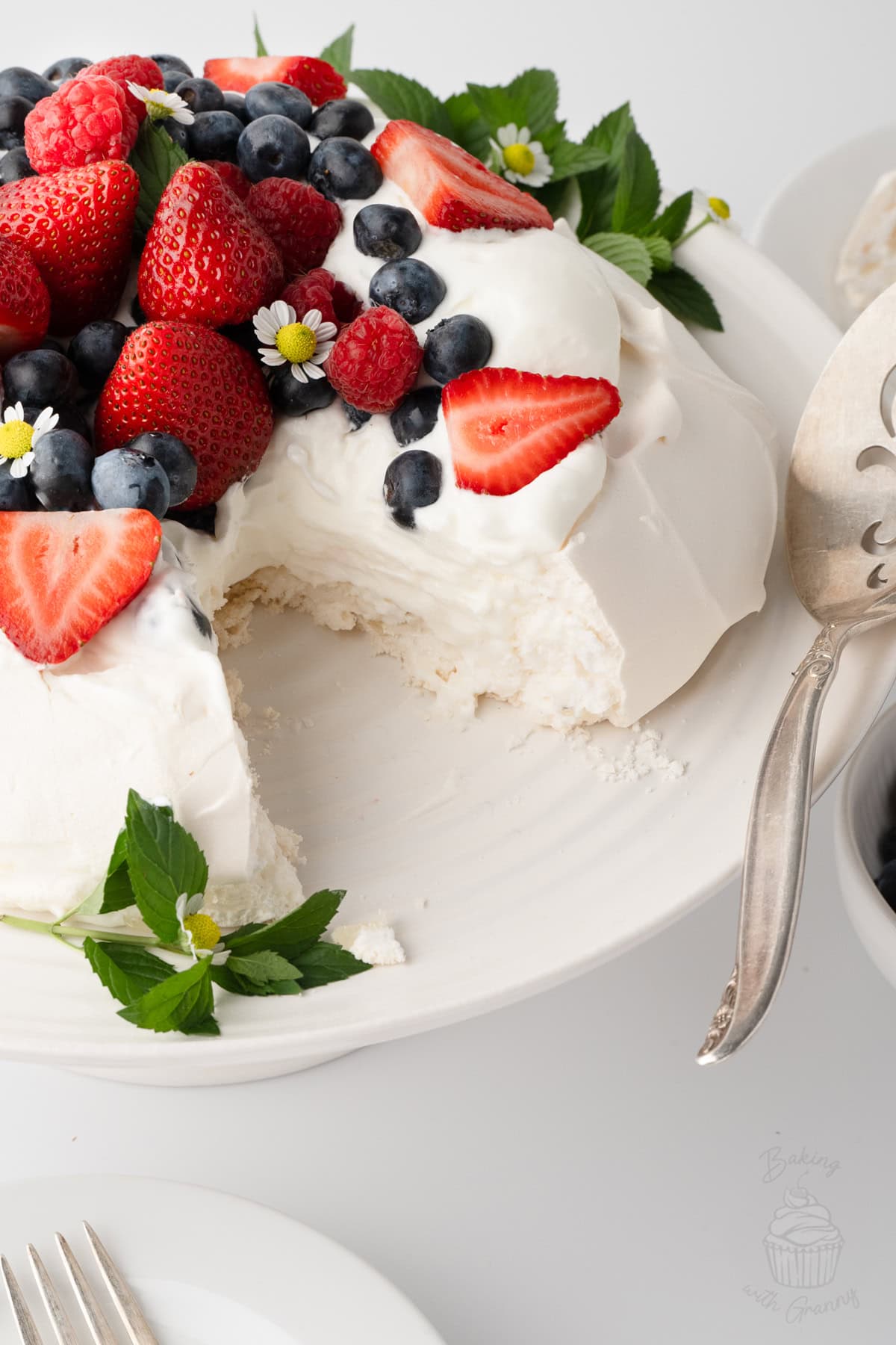 Pavlova topped with whipped cream, strawberries, raspberries and blueberries, with a slice removed.