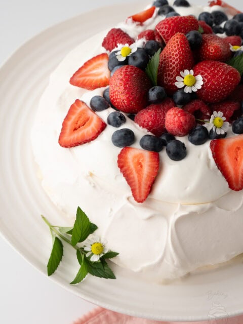 Pavlova topped with whipped cream, strawberries, raspberries, blueberries and small white edible flowers on a plate.