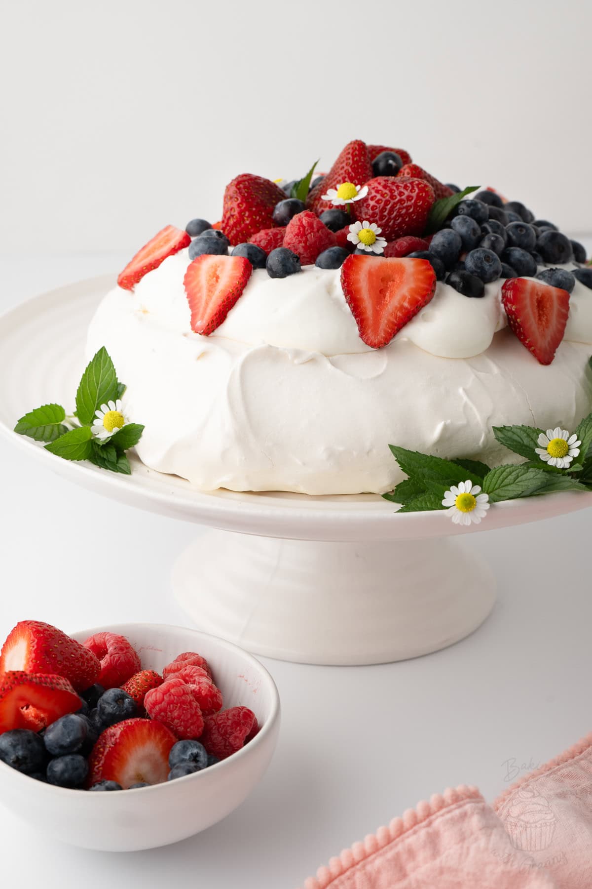 Pavlova topped with whipped cream, strawberries, raspberries and blueberries on a cake stand.