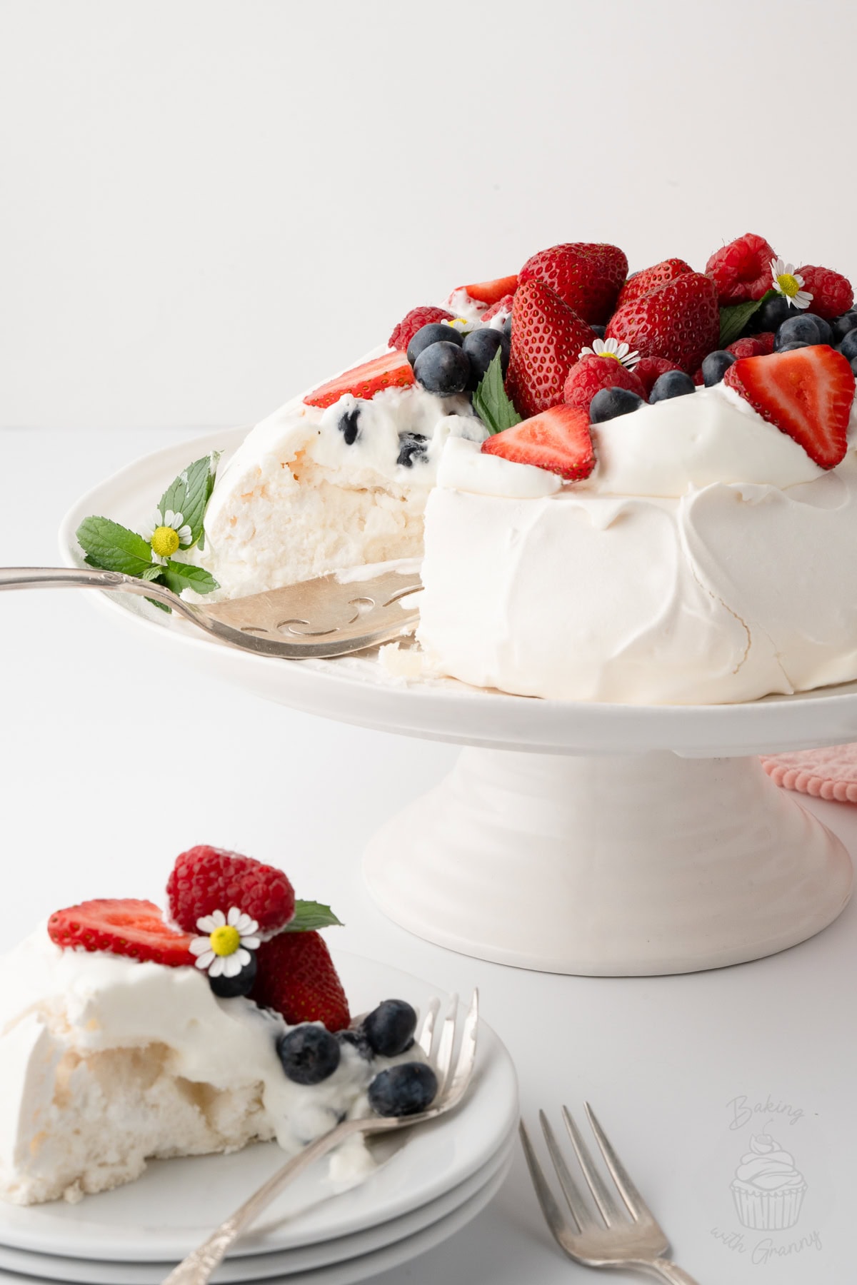 Berry-topped Pavlova on a cake stand with a slice served on a plate in front.