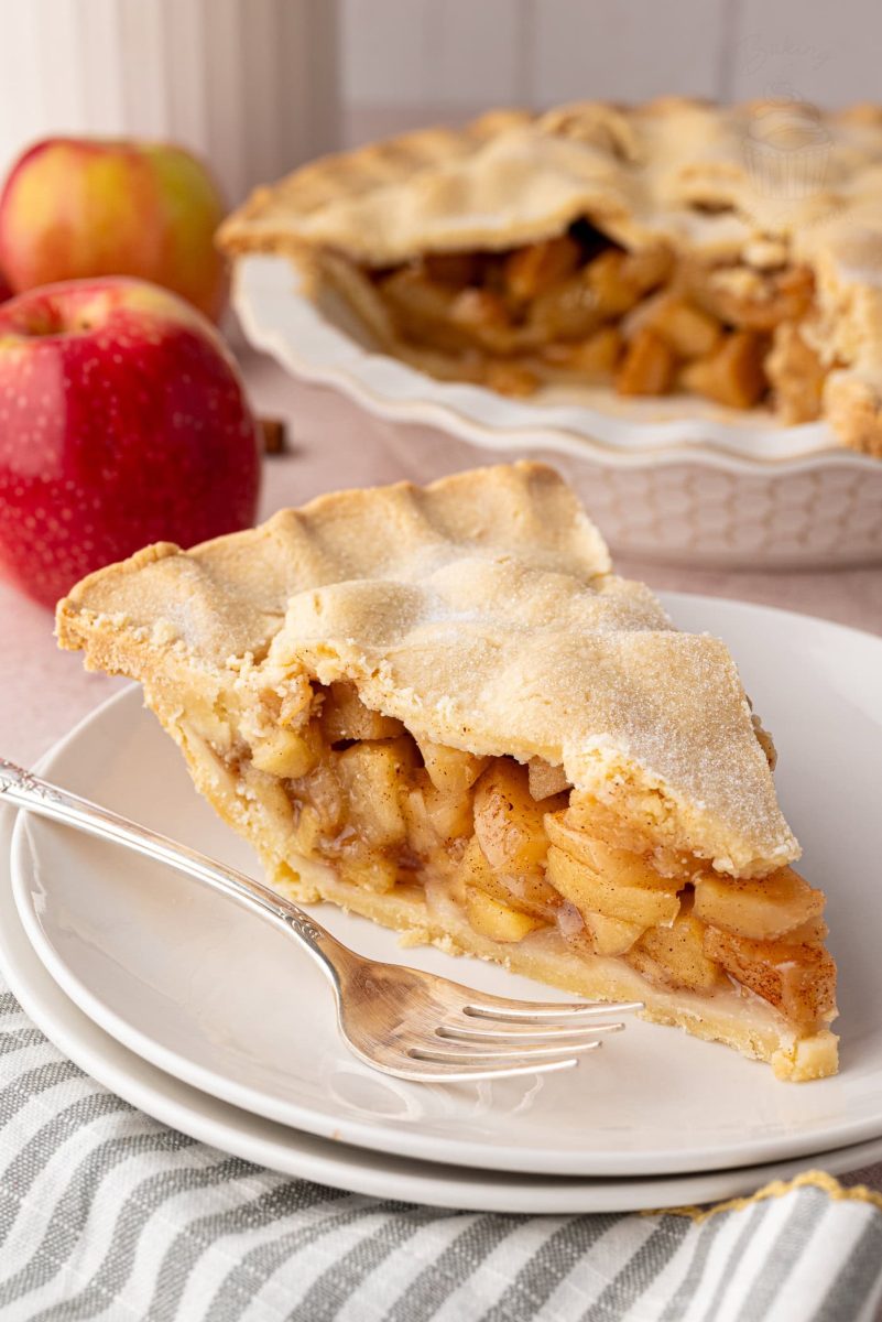 Slice of apple pie on a plate with the remaining pie in the background.