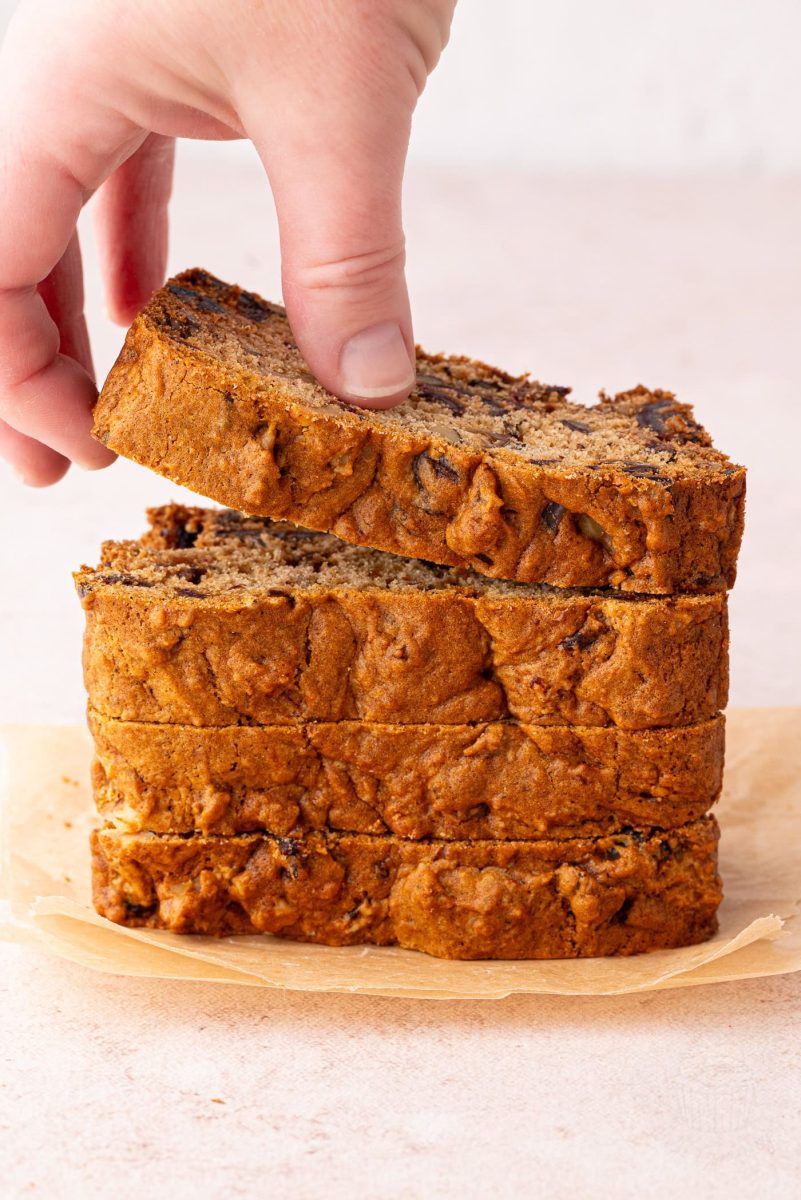 A hand lifting a slice of Date and Walnut Loaf from a stack of four slices on top of parchment paper.